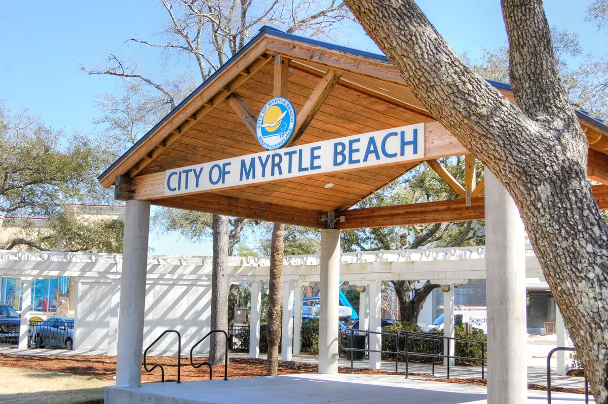 This image features a pavilion structure marked with clear 'CITY OF MYRTLE BEACH' signage, nestled among lush trees. The scene captures a welcoming public space often used for community gatherings, tourist information, and local events, reflecting the area's vibrant culture.