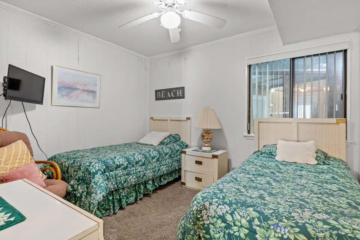 This image depicts the second bedroom with two twin beds adorned with floral bedding, a beach-themed wall art, a lamp with a decorative base, and a small side table between the beds. The layout is bright and inviting, ideal for families or friends sharing the space.