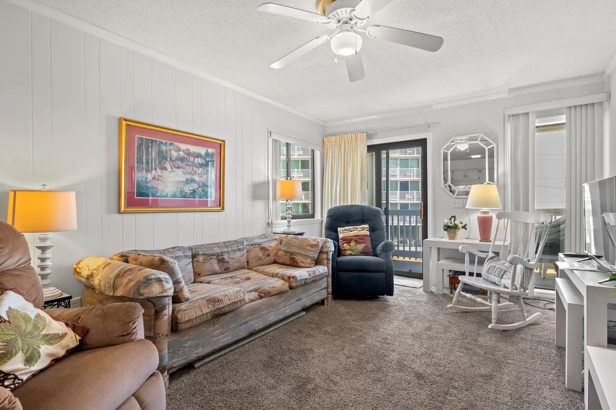 The image showcases a bright and inviting living area with a plush, comfortable sofa, a classic wooden rocking chair, a tasteful decorative painting, a sleek TV unit, and a large window that lets in natural light. The scene is set for relaxing evenings, cozy gatherings, or simply unwinding after a day at the beach.