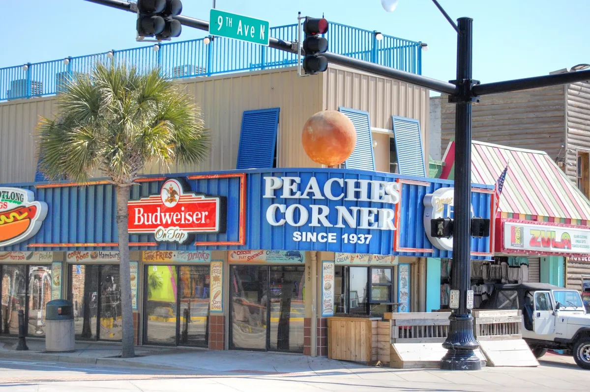 A vibrant streetscape with the iconic Peaches Corner sign, colorful building facades, and palm trees, the scene buzzes with energy. The image highlights local dining options, social spaces, and hints at the lively atmosphere that characterizes the heart of Myrtle Beach.