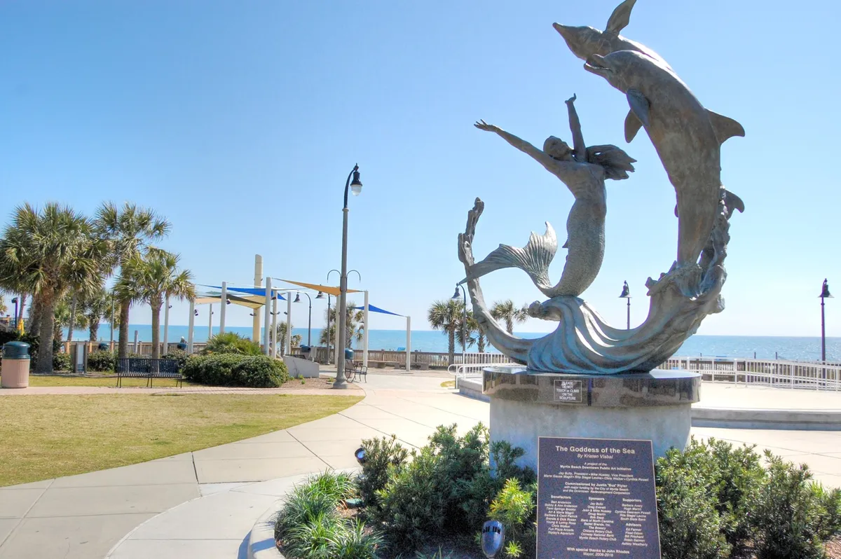 A scenic view along the Myrtle Beach promenade featuring the iconic Goddess of the Sea sculpture, lush palm trees, and the ocean in the background. The uplifting and serene mood highlights the beauty of the coastline at sunset, with inviting spaces for romantic strolls and moments of quiet reflection.