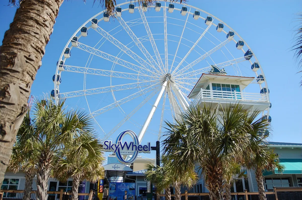 A vibrant exterior shot featuring the Myrtle Beach SkyWheel, with palm trees framing the scene. The festive mood and clear view of the iconic Ferris wheel capture the excitement of nearby attractions, ideal for couples or groups seeking memorable experiences during their getaway.