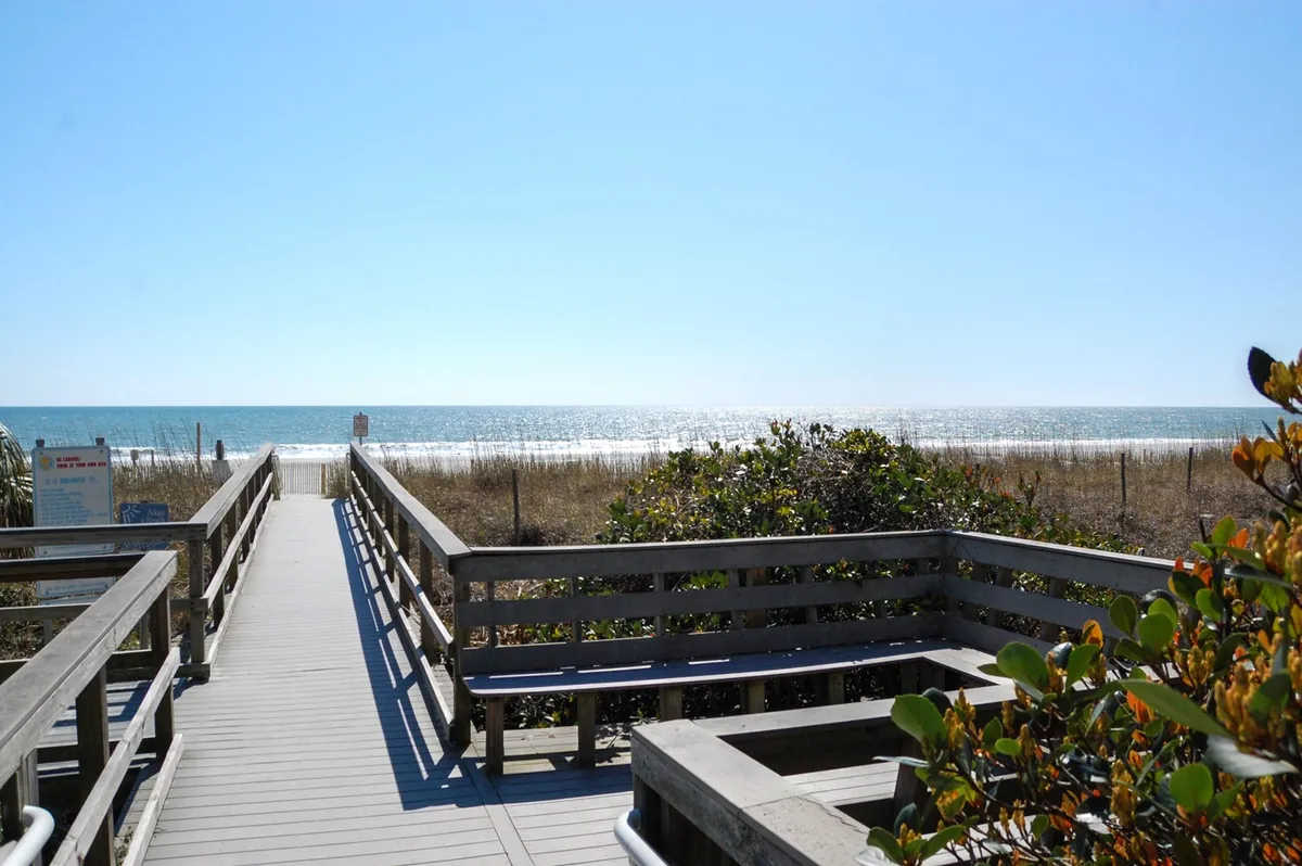 A scenic wooden walkway stretches over dune grass, leading directly to the sandy beach and sparkling ocean beyond. The view is tranquil and inviting, highlighting easy beach access and natural beauty.