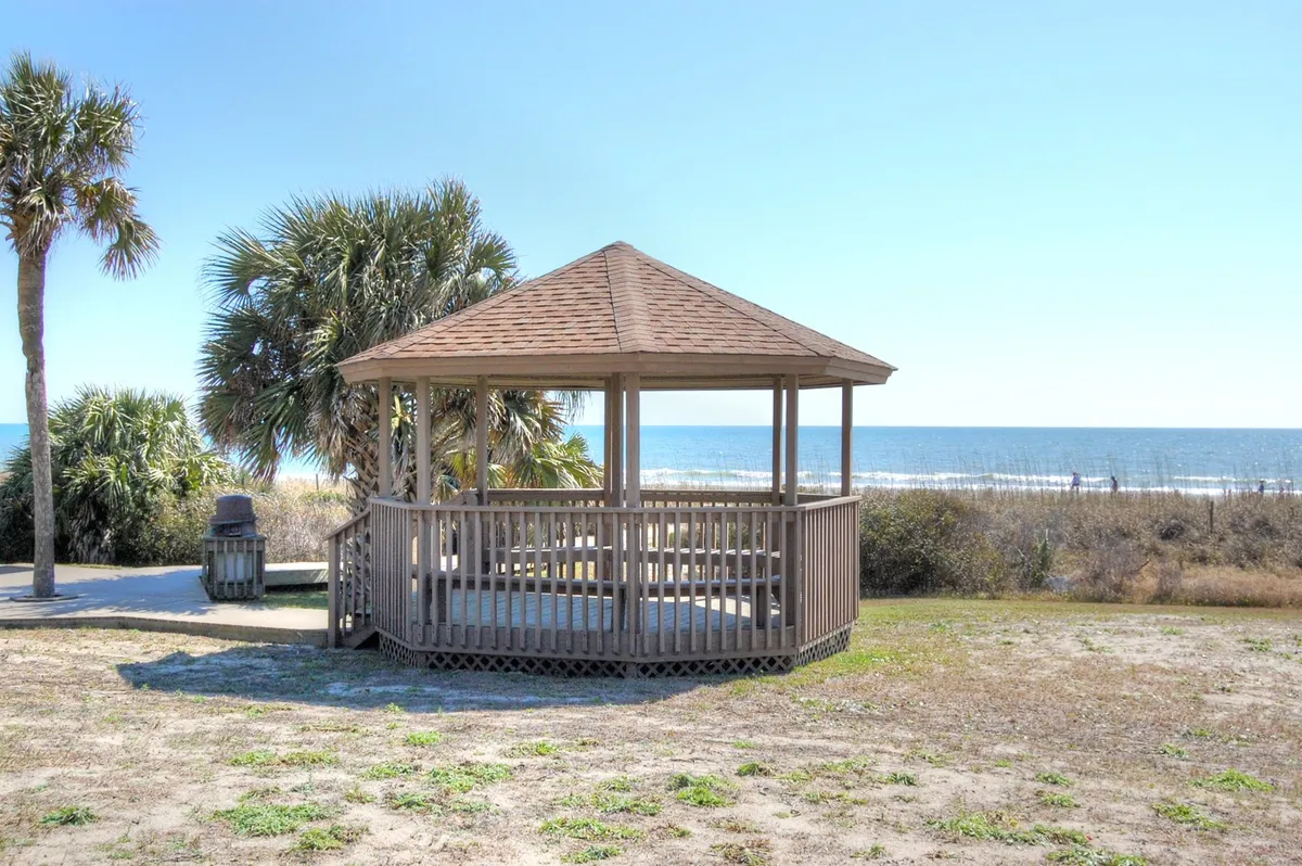 A tranquil outdoor scene featuring a wooden gazebo surrounded by palm trees, with the ocean visible in the background. The setting invites relaxation, with ample space for groups to gather and enjoy the natural coastal environment.