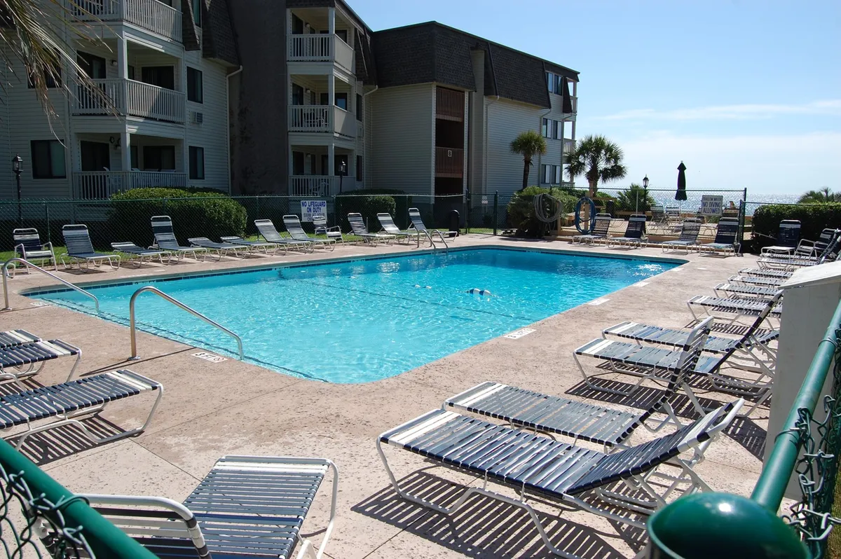 An inviting pool area with numerous lounge chairs, lush greenery, and the building's facade in view. The scene suggests a casual, social setting perfect for sunbathing or gathering with friends throughout the day.