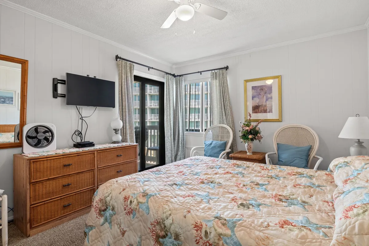 The image shows a tranquil primary bedroom featuring a bed with a floral quilt, a dresser with a mirror, a balcony door letting in natural light, and two wicker chairs arranged for relaxation. Lamps on the nightstands and subtle decor elements contribute to the serene atmosphere.