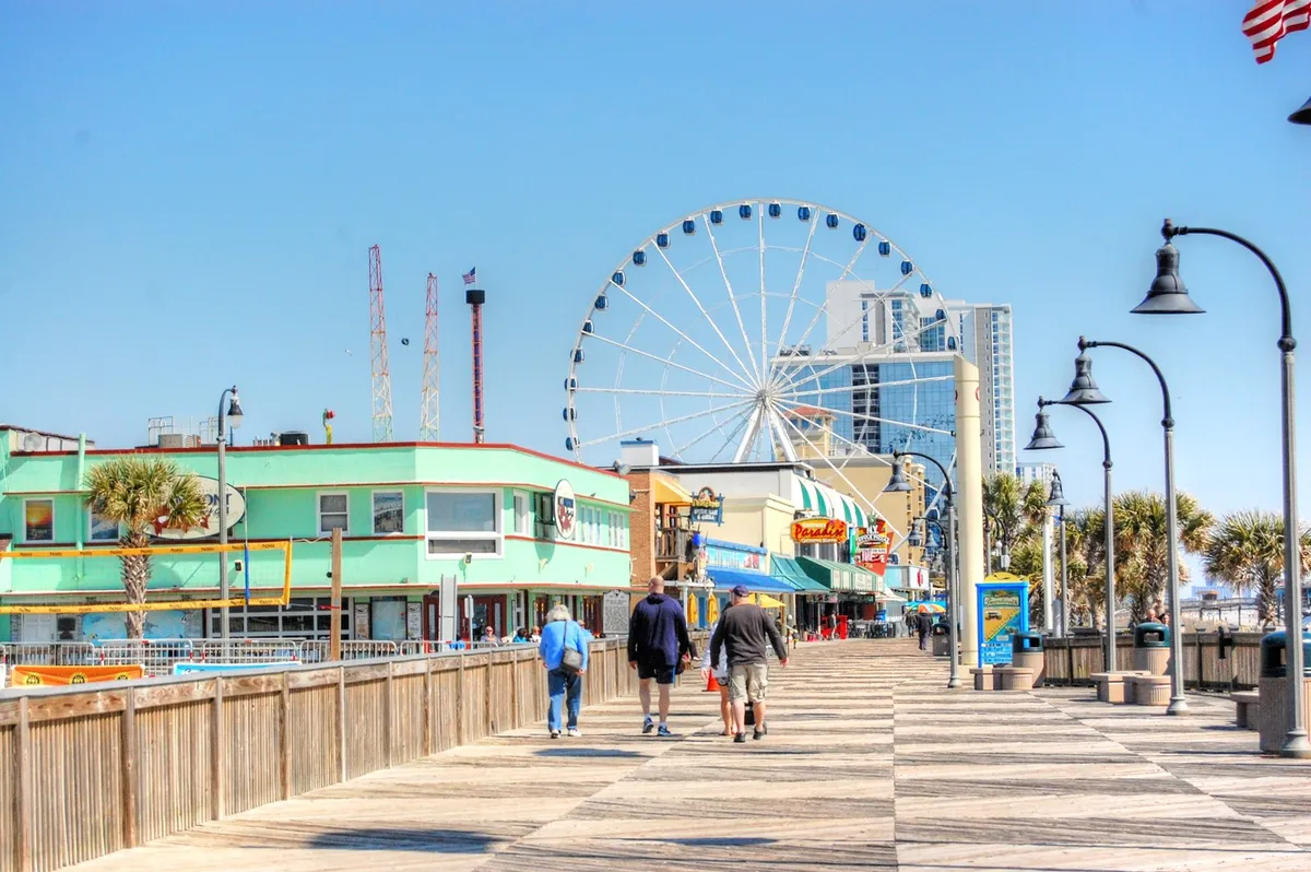 A lively boardwalk scene featuring the iconic Myrtle Beach SkyWheel, colorful buildings, and families strolling along the promenade. The festive atmosphere reflects local attractions and vibrant activity.